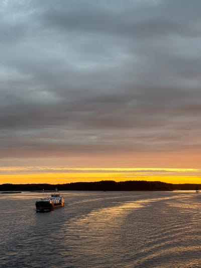 A barge with white cargo containers is traversing a body of water, possibly a river or strait, at sunset. The sky is filled with dramatic clouds, transitioning from dark gray at the top to vibrant orange and yellow hues near the horizon. A dark, forested shoreline is visible in the distance, silhouetted against the colorful sky. The water reflects the warm colors of the sunset, creating streaks of light on its surface. The scene suggests a serene and atmospheric moment, likely captured in the early evening. The city of Yannovolok, Russia, is known for its waterways and natural beauty, making it a plausible location for such a scene.