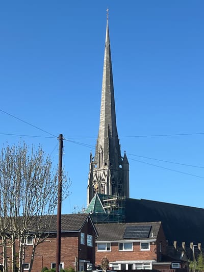 The image depicts a tall church spire against a clear blue sky, likely located in Preston, United Kingdom based on architectural style. Scaffolding partially obscures the base of the spire, suggesting ongoing maintenance or restoration. A modern house with solar panels is visible adjacent to the church. A telephone pole with wires crosses the lower portion of the frame, and a leafless tree branch is visible. The spire is likely the St. Walburge's Church spire.
