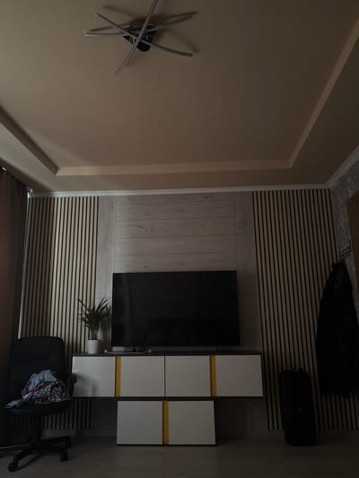 The image depicts a modern living room with a minimalist aesthetic, likely in Częstochowa, Poland. The room features a neutral color palette with a beige ceiling and walls paneled with vertical wooden slats and a horizontal wood-effect panel behind a large flat-screen television. A dark office chair with clothes draped over it sits to the left of the television stand, which is a white unit with yellow accents. A potted plant is placed on top of the television stand, to the left of the television. A tall, black speaker stands to the right of the television stand, and a coat is hanging on a wall hook on the right side of the frame. A unique ceiling light fixture with multiple metallic arms is visible at the top of the image. The lighting is dim, creating shadows and highlighting the textures of the room. There are no people visible in the image, and no explicit actions are taking place.