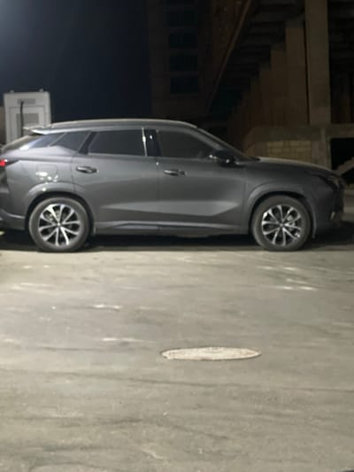 A gray SUV is parked on a paved surface at night, possibly an urban area. The vehicle is seen from its side profile, showing both the front and rear wheels. The wheels have a distinctive multi-spoke design. In the background, there are structures that suggest an industrial or construction site, with visible concrete pillars and beams. The lighting indicates it is nighttime. The image was taken in Sumgayit, Azerbaijan.