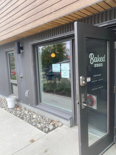 The exterior of a business called "Baked CAFE" is shown in Whitehorse, Canada. A dark gray door is ajar, revealing a glass entrance. The door has a silver digital lock and a silver handle. On the glass of the door, the business name "Baked CAFE" is written in white lettering, along with the cafe hours: Monday to Friday, 8:00 AM to 4:00 PM, and Saturday, 9:00 AM to 3:00 PM. A "WARNING" sticker is visible on the door.
To the left of the door is a large window with signage displayed inside. One sign states "WE WILL BE CLOSED ON Canada Day." Another sign reads "UBE SOFT SERVE ICE CREAM." A yellow circular light fixture is visible through the window. The building has corrugated metal siding and a wooden soffit. A black outdoor light fixture is mounted to the siding. A white planter with a plant is positioned near the window. The ground in front of the building is paved with concrete and bordered by rocks and some fallen leaves.
