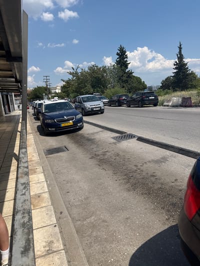 The image shows a street scene in Menemeni Municipal Unit, Greece, on a sunny day with scattered clouds. In the foreground, a tiled sidewalk runs along the left edge of the frame, with a metal railing casting a shadow. A person's lower legs, wearing socks and athletic shoes, are visible on the sidewalk. The street is paved with asphalt and lined with parked cars, primarily taxis. A blue Skoda Octavia taxi with a yellow license plate and "TAXI" signage is prominently featured in the middle ground. To the right of the street, a grassy area with trees and bushes separates the road from a slightly elevated background featuring buildings and utility poles. Several concrete barriers and traffic control devices are present along the roadside. The overall impression is that of a typical urban street with vehicle traffic and pedestrian infrastructure.