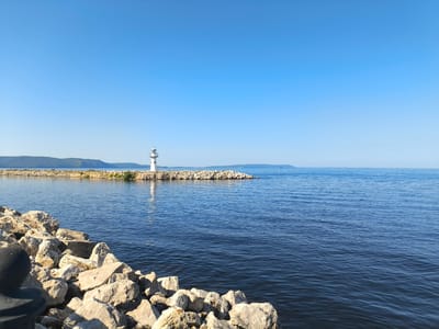 The image depicts a clear, sunny day at the waterfront in Volzhskiy Utes, Russia. In the foreground, large, light-colored rocks are piled up along the edge of the water. The dark blue water is calm with small ripples. In the middle ground, a man-made breakwater, also constructed of rocks, extends into the water. At the end of the breakwater stands a white lighthouse with a green-capped lantern room. The breakwater is topped with some sparse vegetation. In the background, a long, tree-covered shoreline stretches across the horizon under a bright blue sky. The distant water is a lighter shade of blue and a small white boat is visible on the water.