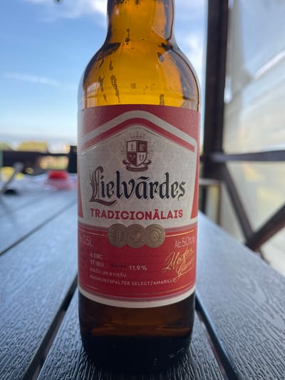 An amber-colored glass bottle of Lielvārdes Tradicionālais beer lies horizontally on a dark, textured wooden table. The bottle is about two-thirds full, and condensation with small bubbles clings to the inside glass, indicating a cold beverage. The main label, predominantly red and white with gold accents, is clearly visible.
In the background, a bright, clear blue sky is visible on the lower left, while a translucent white corrugated roof structure, supported by dark beams, spans the upper background, suggesting an outdoor or semi-outdoor setting like a patio or open-air restaurant. The lighting is bright, indicating daytime.
Visible text on the label includes "Lielvārdes TRADICIONĀLAIS", "0.5L", "Alc. 5.0%", "6 EBC", "17 IBU", "BLĪVUMS: 11,9%", and "MIEŽU UN KVIEŠU" (Barley and Wheat). The hop varieties "MAGNUM/SPALTER SELECT/AMARILLO" are also listed. Other phrases like "DARITS ALUS" (Brewed Beer) and "LABO LAIKA" are present. The image captures a static moment, with no human activity taking place around the bottle, which rests casually on the table in what appears to be Zaķumuiža, Latvia.