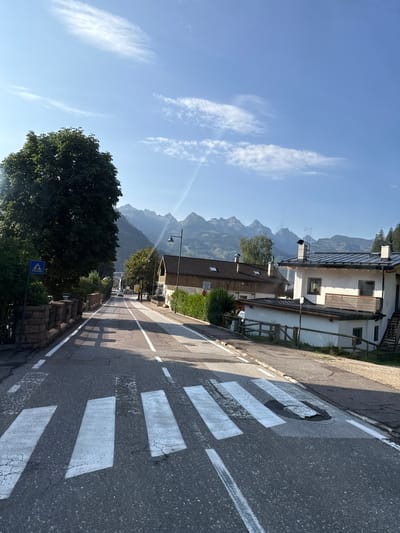 The image depicts a street scene in Predazzo, Italy, on a clear day. The foreground features a pedestrian crossing with prominent white stripes on asphalt. To the left of the road, a large, dark green tree casts a shadow on the asphalt. A stone wall borders the left side of the road, with a blue pedestrian crossing sign visible on a metal post. Further down the road, houses with traditional architecture and pitched roofs are visible, some with visible chimneys. In the background, a mountain range with jagged peaks stretches across the horizon under a bright blue sky with wispy white clouds. The road appears to be a two-lane street with white lane markings. Sunlight illuminates the scene, creating distinct shadows from the trees and buildings.