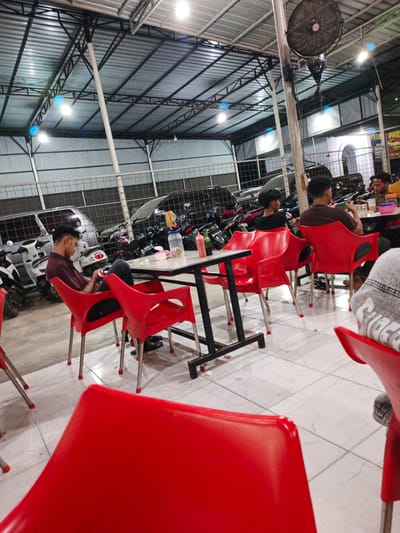 The image captures an indoor dining area, likely a food court or open-air restaurant, with a corrugated metal roof supported by a metal framework. Several bright fluorescent lights illuminate the space. The floor is tiled with a white and grey marble pattern. In the foreground, a bright red chair is prominently featured, partially obscuring the view of the rest of the scene.
The main subjects are several people seated at tables. A young man in a maroon shirt is seated alone in a red chair, looking down at his phone. He is positioned near a table with a white surface, on which rests a bottle of red sauce and a jar of what appears to be food. Further back, two men are seated at another table, engaged in conversation. One man wears a brown shirt and gestures with his hand, while the other wears a black shirt. A third person, wearing a yellow shirt, is partially visible at this table.
Various objects are scattered around the seating area, including red plastic chairs with metal legs, a metal table with a black frame, and several motorcycles parked in the background. The motorcycles suggest this may be a location where patrons arrive on their bikes. Behind the people and motorcycles, a chain-link fence is visible, and beyond that, structures that appear to be buildings with large windows.
A large industrial fan is mounted on a pole near the center of the image. Visible text on a sign in the background reads "PUTRI BOOK". The overall atmosphere appears casual and relaxed, with people dining or socializing. The lighting suggests it could be evening or a dimly lit indoor space during the day. The location context indicates this scene is in Cotpuuk, Indonesia.