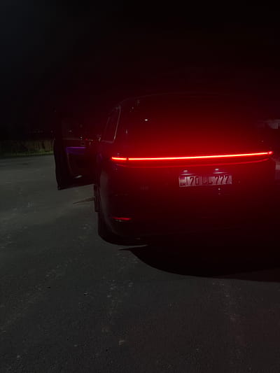A dark-colored car, possibly black, is parked on a dark surface, likely asphalt, in Mkhchyan, Armenia. The car's taillight is illuminated as a single bright red vertical line. The license plate reads "AM 70LL777". The driver's side door appears to be open, with a purple object visible inside. The surrounding environment is dark, with some faint illumination suggesting the presence of vegetation in the background.