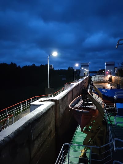 This image depicts a canal lock in the town of Devyatiny, Russia, at dusk. The scene is dominated by a dark, cloudy sky with dramatic lighting from street lamps illuminating the lock walls and a nearby boat. The lock is a substantial concrete structure, with a railing running along its edge. Inside the lock, two barges are positioned, one blue and one white. A smaller orange lifeboat is attached to the side of one of the barges. The surrounding environment appears to be a forested area, visible as a dark silhouette against the twilight sky. The overall atmosphere is quiet and industrial, illuminated by artificial light against the natural darkening of the evening.