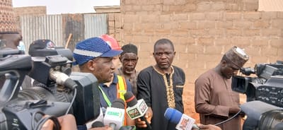 An outdoor scene in Rigasa, Nigeria, depicts a media event with five adult males prominently featured in the mid-ground and foreground. In the foreground left, a man wearing a blue and white striped cap over a dark blue shirt and an orange and yellow reflective safety vest is speaking into multiple microphones, one labeled "KSMC" and another "ARISE NEWS." To his immediate right, partially obscured, is an individual in a red hard hat. Behind the speaker, towards the center, an older man with a dark cap and patterned shirt looks forward intently. Adjacent to him, a man in a dark kaftan with gold embroidery also looks directly forward. To the right, a man in a brown traditional outfit and a patterned cap is looking downwards, possibly adjusting a black professional video camera. Multiple other professional video cameras are positioned in the foreground and mid-ground, with one camera clearly showing a silver XLR cable connected to its side. The background consists of a light brown, exposed brick wall and a section of corrugated metal fencing, indicating a raw or developing construction environment. The ground appears to be reddish-brown earth.