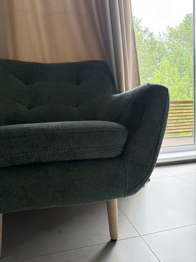 A dark green textured fabric sofa is positioned in a room with a large window. Through the window, lush green trees and a wooden railing are visible. Light-colored curtains hang on the right side of the window, partially obscuring the view. The sofa has a tufted back and a rounded armrest. It rests on light-colored wooden legs. The floor is tiled with large, light grey tiles. The scene appears to be set in a residential area, likely a living room, in Kamyennoye, Russia, based on the provided context.