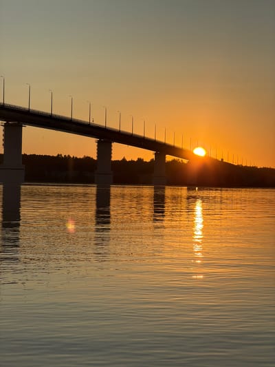 A long, elevated highway bridge spans across a wide river, silhouetted against a warm, gradient sunset sky. The sun, a bright orb, is partially obscured by the bridge, casting a golden glow and a shimmering reflection on the water's surface. The bridge has a railing and is lined with regularly spaced lampposts. On the far bank, a dense line of dark trees forms a horizon. The water exhibits gentle ripples, creating a textured surface that reflects the vibrant colors of the sky. The overall scene is peaceful and serene, capturing the beauty of a sunset over a river and its accompanying infrastructure in the city of Kambarka, Russia.