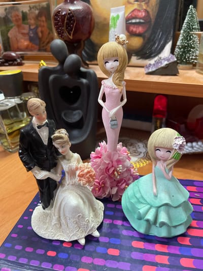 The image depicts a collection of decorative figurines arranged on a wooden surface. In the foreground, a wedding cake topper features a groom in a black tuxedo and a bride in a white lace gown holding a bouquet of pink roses. To the right, a figurine of a young girl in a teal dress with layered ruffles stands beside a larger figurine of a woman in a pink dress and a small purse, adorned with a crown. Behind these, several other objects are visible, including a black sculpture of embracing figures with a heart cutout, a framed family photograph, a decorative pomegranate, a small Christmas tree, and bottles of perfume or cosmetic products. The overall scene suggests a display of sentimental or decorative items. The specific location of Pokoinoe, Russia, is not discernible from the image itself.