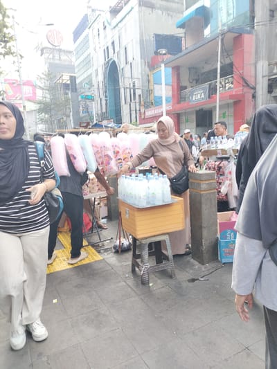The image depicts a street scene in Central Jakarta, Indonesia, likely a commercial area given the buildings and pedestrian activity. In the foreground, a woman wearing a black and white striped shirt, tan pants, and white sneakers stands on a tiled sidewalk. She is facing left, with her left hand extended and holding a smartphone. To her left, another woman is partially visible, wearing a black hijab and a striped shirt.
Behind the foreground woman, a vendor is selling cotton candy, with several pink and blue cotton candy packages hanging from a display. Next to the cotton candy display, a woman in a tan hijab and dress is attending to a stack of bottled water. She is reaching towards the bottles. In the background, there are several buildings, one with a large red sign that appears to be an advertisement. Other individuals are visible in the midground, including a man wearing a black shirt and dark pants, and others who are less clearly defined. The environment is an urban street with a sidewalk, suggesting a marketplace or a busy thoroughfare. There are some trash bags and a spill on the ground near the bottled water display.