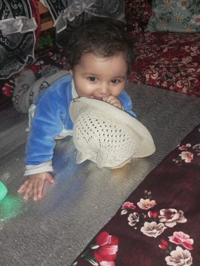 A baby lies prone on a mat. The baby, wearing a blue velvet top and grey pants, is partially covering a beige straw hat. The mat has a silver, textured surface with a floral-patterned cloth in the lower right. The background shows fabrics and a pillow with a daisy pattern. The photograph may have been taken in Galaosiyo, Uzbekistan.