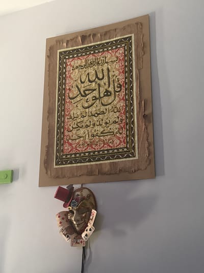 A framed piece of calligraphy, written in Arabic script, is hanging on a light-colored wall. The calligraphy is on a textured background, with ornate patterns in red and gold. The frame itself appears to be made of a rough, aged material, possibly papyrus. Below the calligraphy, a decorative mask is hanging. The mask is elaborate, with gold and intricate designs, and is adorned with a small red hat and playing cards. The scene is indoors, likely in a home or a shop, and there is a hint of another object on the left side of the image which is green. The lighting suggests it is daytime. There are no people visible in the image. The image does not provide any specific information about the city of Rimini, Italy.