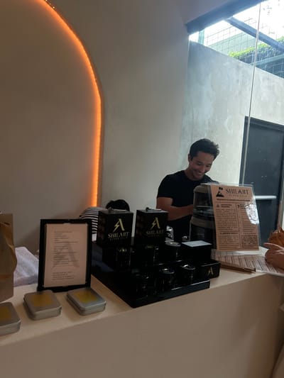 The image shows a man standing behind a counter with a product display, located at coordinates 9.952, -84.196 in Pozos, Costa Rica. The man is wearing a black shirt and smiling. He appears to be interacting with a customer, who is not visible in the image. On the counter, a display of small black boxes with a gold "A" logo on top, labeled "SHILAIT Black Gold," is visible. Another sign, also labeled "SHILAIT Black Gold," provides information about the product. To the left of the display, a black frame contains a menu, and two small silver boxes with gold accents are visible. On the table is a white towel and a paper bag. The background shows an orange light, a wall, and a window with a view of greenery.