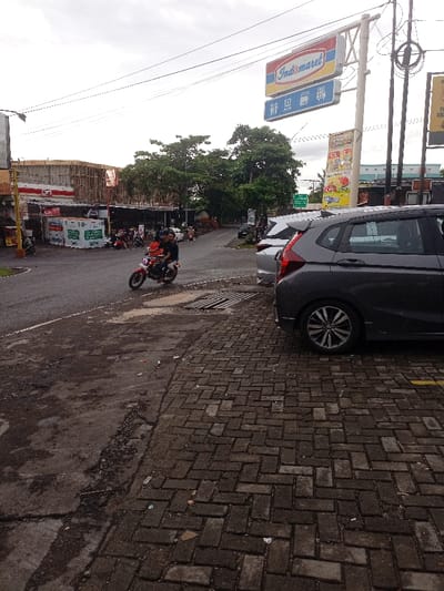 The image shows a street scene in Kecamatan Caringin, Indonesia (latitude: -7.523, longitude: 107.485).  A man and a woman are riding a red and black motorbike on a paved road with a brick sidewalk to the right. The motorbike is moving to the left. There are two cars parked on the right side of the street, a grey Honda Jazz and a silver SUV. Both cars are facing to the left. In the background, there are a number of buildings and trees. A sign for a supermarket is visible on the right side of the image. There is an overhead power line running across the image, as well as a streetlight. The sidewalk is made of brick and appears to be in disrepair. The road is paved and appears to be in good condition. There is a drainage grate in the middle of the road. The weather is overcast and there are no clouds visible. The street is empty except for the motorbike and the cars.  There is a yellow line on the road. There is a small amount of debris visible on the sidewalk.  The image appears to have been taken from the perspective of a person standing on the sidewalk.