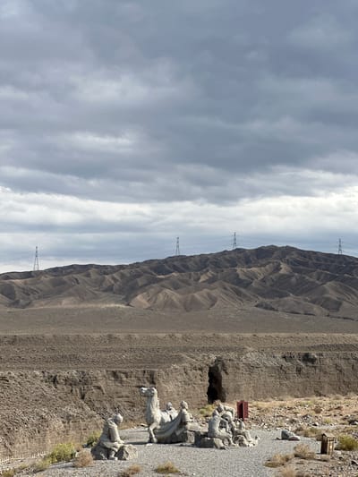 The image depicts a desolate, mountainous landscape with a cloudy sky. In the foreground, to the right, there are several stone sculptures of people and horses, arranged on a gravel path. The figures appear to be in a historical or military context, possibly representing soldiers or warriors. One sculpture shows a person riding a horse, while others depict figures standing or in action. Behind the sculptures, the terrain is rocky and arid, with sparse vegetation.
The background is dominated by a large, eroded mountain range with sharp, jagged ridges. Several tall, modern-looking transmission towers with power lines are visible against the sky and the mountain slopes, suggesting the presence of infrastructure in this remote area.
The scene appears to be outdoors, under a sky filled with layered, greyish-white clouds, indicating overcast or partly cloudy weather. The lighting suggests it is daytime, though the exact time is difficult to ascertain due to the cloud cover. The overall atmosphere is one of ruggedness and historical significance, with the sculptures providing a focal point against the natural backdrop. The location is identified as Jiayuguan, China, which is known for its historical fortifications and surrounding desert landscapes. There is no visible text in the image.