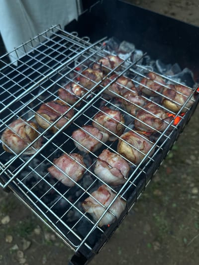 A metal grill basket is filled with food items wrapped in bacon and being cooked over hot coals. The grill basket is angled, showing multiple rows of bacon-wrapped food, some appearing to be small potatoes or dense vegetables. The bacon is beginning to crisp and brown, with some pieces showing rendered fat. The coals underneath are glowing red, indicating intense heat. Smoke is rising from the grill. The setting appears to be outdoors, with dark soil or dirt visible in the background, interspersed with some green vegetation. A portion of a black grill body is visible to the left of the basket. The lighting suggests it could be daytime, with diffused light. There is no visible text in the image. The overall impression is a casual outdoor cooking scene, likely a barbecue.