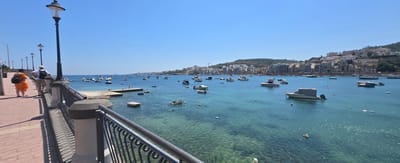 The scene is a bright, sunny day in Saint Paul's Bay, Malta. The view is of the Mediterranean Sea from a waterfront walkway with a metal railing. The sea is clear and turquoise in color, with many small boats and some larger boats dotted across the bay. The shoreline is visible in the background, with buildings and hills rising up from the coast. Two people are walking on the walkway, one wearing an orange dress and carrying a backpack, and the other wearing a white shirt and khaki shorts, also with a backpack. Several streetlights are visible along the walkway.