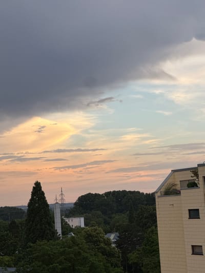 This image captures a sky filled with dramatic clouds, transitioning from dark gray on the left to lighter hues of blue and pastel orange towards the right. A prominent, lighter band of cloud in a diamond-like shape is illuminated by sunlight. Below the sky, a dense line of trees forms the horizon, with a tall, slender evergreen tree standing out on the left. In the background, industrial structures are visible, including a tall, multi-poled electrical tower and a white rectangular building. On the right side of the frame, a modern, light-colored building with several windows and a balcony with greenery is visible, extending into the lower right corner of the image. The scene appears to be a view from a window or elevated vantage point in Sindelfingen, Germany, likely during dusk or dawn, given the sky's coloration. There are no people visible in the image.