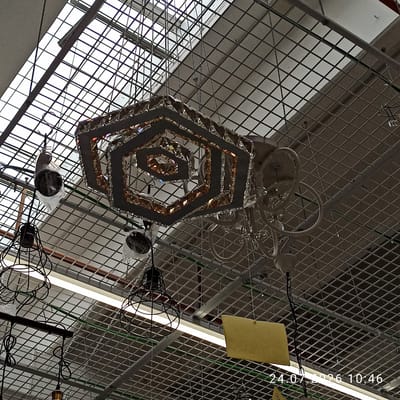 The image shows a collection of chandeliers and pendant lights hanging from a metal grid ceiling in what appears to be a retail store. The main chandelier, centrally located, is hexagonal and adorned with numerous faceted crystals, reflecting light and creating a prismatic effect. Several other pendant lights are visible, including some with wire cages and others with reflective shades. The ceiling structure is made of interconnected metal bars, supporting the lights and also featuring some fluorescent lighting fixtures. The overall scene suggests a display of lighting fixtures, possibly in a home improvement store or a specialized lighting showroom. The timestamp "24.07.2026 10:46" is visible in the lower right corner of the image, indicating when the photograph was taken. The location is stated as Modugno, Italy. There are no people visible in the image.