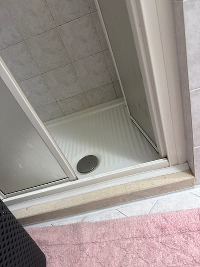 An interior shot of a bathroom shows a white shower stall with a textured floor and a metallic drain cover. The shower stall has a clear glass door. The floor outside the shower is tiled with light gray tiles. A portion of a pink rug is visible in the foreground, and a dark gray rack is partially visible on the left side. The scene appears to be in Villa Pasquali, Italy.