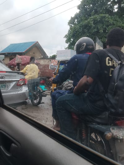 This media file captures a scene of traffic congestion in Alagbede, Nigeria, under an overcast sky, suggesting either early morning or a rainy day. The primary subjects are two men on a motorcycle and a person in a car.

The person in the car is partially visible through the window, and appears to be looking out. The two men on the motorcycle are the central focus. The rider wears a dark blue outfit and a black helmet with a silver design. The passenger, seated behind the rider, wears a black t-shirt with gold lettering that reads "GA" and partially visible "D". He also has a black backpack slung over his shoulder. The motorcycle itself is dark-colored, with a visible rear shock absorber.

In the background, several elements suggest an urban or semi-urban environment. A silver car with a license plate reading "LAGOS -829FU" is parked alongside the motorcycle. Further back, a building with a distinctive blue-tiled roof and green trim is visible. Red umbrellas are set up, possibly for vendors. Signage for "POS DATA" and "EXO TOKEN" is present, indicating commercial activity. Another sign for "ST. PAUL'S CATHOLIC" and "EBUTE-METIN" provides further location context. The ground is wet, reinforcing the idea of recent or ongoing rain, and the overall atmosphere is one of a typical busy day in Nigerian traffic.