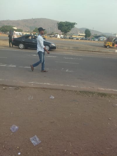 The image shows a man walking on a paved road in Maraba, Nigeria. The man is wearing a white long-sleeved shirt, blue jeans, and a black cap. He is walking away from the camera. There is a black car parked on the side of the road. There are other vehicles in the background, including a yellow three-wheeled vehicle. The environment is urban, with a dirt shoulder on the right side of the road. There are several pieces of litter on the dirt shoulder. The scene is a typical example of a busy urban environment.  The coordinates provided, latitude: 9.024, longitude: 7.573, place the image in Maraba, Nigeria.
