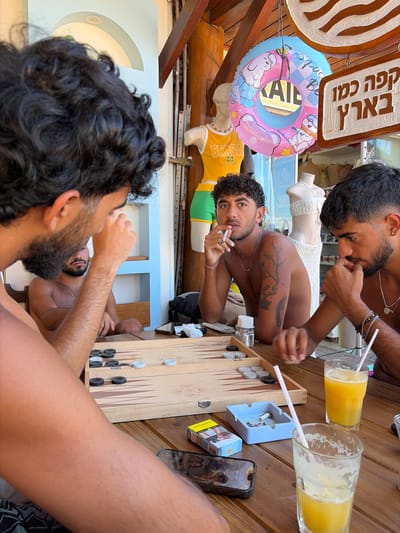 The image shows three men seated around a backgammon board on a wooden table. The setting appears to be outdoors in Cairu, Brazil, indicated by a "BRASIL" t-shirt hanging in the background. Two of the men have dark beards, and one of them is holding a cigarette. There are two glasses of orange juice with straws on the table, along with an ashtray, a pack of cigarettes, and a smartphone. A colorful inflatable pool float is hanging nearby. The backgammon game is in progress.