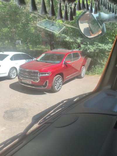 The image shows a red GMC Acadia parked on a paved surface, possibly a street or parking lot, in Alchevsk, Ukraine. The car is a four-door SUV with a prominent chrome grille and alloy wheels. A white sedan is parked to the left of the GMC. The scene is viewed from inside a vehicle, with the windshield wipers and dashboard visible in the foreground. Trees and foliage are visible in the background, suggesting an outdoor setting. There are no people visible in the image. The weather appears to be sunny.