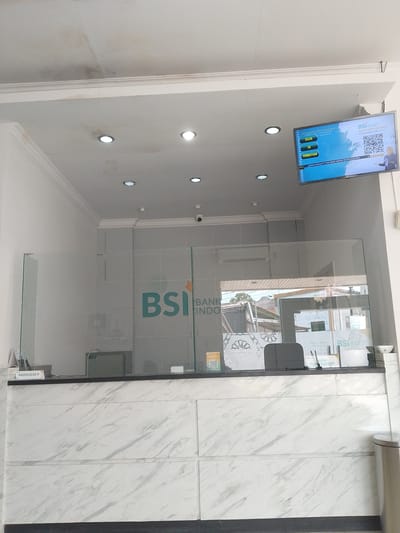 The image shows the interior of a BSI Bank Indonesia branch, possibly located in Manetunong, Indonesia. A teller counter with a marble-like facade and a glass barrier spans the foreground. The BSI logo is visible on the barrier. A monitor displays information, including queues for tellers, CS, and PEMBIAYAAN. The counter has computer equipment, stationery, and a nameplate reading "NADIA ELSA P". The ceiling has recessed lighting. Outside, the reflection shows houses and power lines. A CCTV camera is also seen.