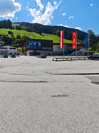 A sun-drenched scene unfolds in Filzen, Austria, featuring a modern building facade adorned with flags and a large mural. The building, likely a transportation hub or tourist facility, displays prominent signage indicating "WAGSTATTBAN" and "10 EUR - Comfortline." Three vertical flags, representing Austria, a red flag with a white goat emblem, and the European Union, flutter in the breeze. Behind the building, a lush green hillside rises, dotted with trees and a ski lift, hinting at winter sports activities. Several cars are parked in the foreground on an asphalt lot, their presence suggesting a public access area. The sky is a brilliant blue, punctuated by fluffy white clouds.