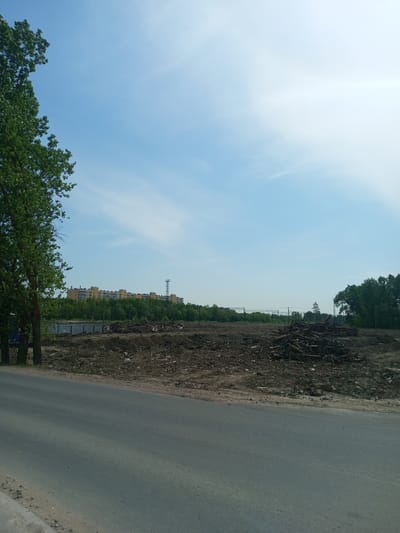 The image depicts a landscape in Kommunar, Russia. A paved road is in the foreground. Beyond the road, the scene transitions to a dirt and debris-covered area. Piles of wood debris, possibly from felled trees, are visible in the middle ground. Trees are present on both the left and right sides of the image. In the distance, buildings can be seen. The sky is blue with scattered clouds.