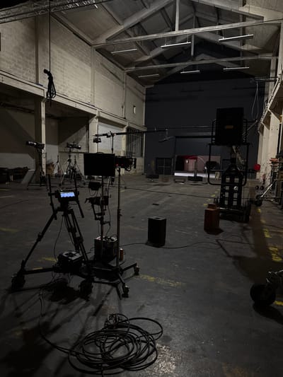 The image depicts the interior of a large, high-ceilinged industrial-style space, identified as a film or photography studio in Ivry-sur-Seine, France. The environment features exposed concrete block walls, a concrete floor, and a pitched roof supported by concrete beams from which linear fluorescent light fixtures are suspended. The overall lighting is dim and artificial, creating pronounced shadows.

In the foreground, a professional video camera system is mounted on a tripod equipped with wheels, positioned over a coil of black cables on the dark concrete floor. A monitor is attached to the camera rig. Immediately to its right, a rolling production cart supports another monitor and various electronic components. A black rectangular speaker is placed on the floor to the right of this cart.

Further into the mid-ground, multiple studio lighting fixtures on stands are arranged on the left side, some with diffusers. A heavy black rope hangs vertically from an overhead beam on the left. In the background, a large dark grey or black backdrop wall conceals a roll-up garage door, through which an illuminated exterior space is visible. To the right, a tall, heavy-duty rolling stand or cart supports a large black object, possibly another speaker or light fixture. An orange canister rests on the floor next to this cart. Scattered cables are visible across the floor. No individuals are present in the scene.