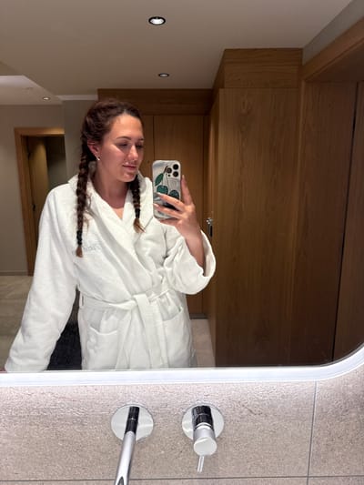 A woman with braided hair, wearing a white bathrobe, stands in a bathroom and takes a selfie in a mirror. The bathroom features wood-paneled walls and beige tiled surfaces around the sink. The woman is holding a smartphone with a cherry-patterned case. The location is identified as a bathroom in Mayrhofen, Austria.