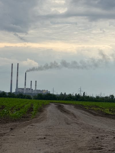 The image presents a landscape juxtaposing agricultural land with an industrial complex, set under an overcast sky in Zanky, Ukraine. In the foreground, a textured, light brown dirt path, appearing somewhat damp, curves gently and recedes into the distance. This path is bordered by a vibrant green field, likely cultivated crops, which forms a dense line across the lower left and center of the frame.
Behind the green field, a line of darker green trees separates the foreground from the industrial elements in the midground and background. The industrial complex is dominated by several tall, slender chimneys, painted with alternating white and red bands. One of these chimneys is actively releasing a thick, dark grey plume of smoke that rises into the sky, indicating ongoing operations and industrial activity. Various other structures of the industrial plant are visible behind the chimneys. Further across the landscape, a series of electricity transmission towers (pylons) can be seen.
The sky is largely overcast, filled with varying shades of grey and white clouds. A distinct, brighter vertical band of light cuts through the clouds on the right side of the image, suggesting either diffused sunlight breaking through heavy cloud cover or the light of an early morning or late afternoon sun. There are no people visible in the image, nor is any text present. The scene effectively captures the coexistence and contrast between nature and heavy industry.