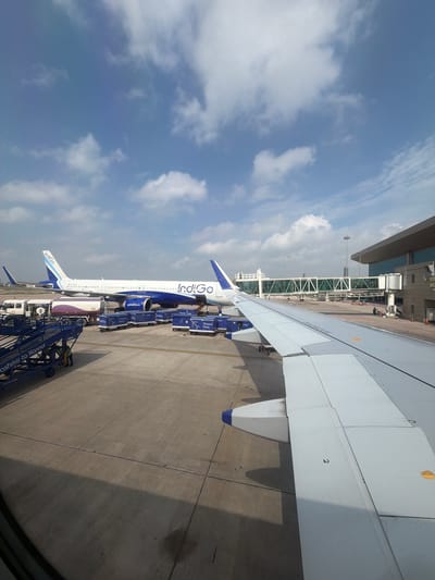 An aerial view captures an Indigo airline plane parked on the tarmac at an airport, possibly in the vicinity of Vikram Nagar, India, under a partly cloudy blue sky. The aircraft, predominantly white with blue accents, is labeled with "Indigo" on its fuselage and tail. Several ground support vehicles and equipment, including luggage carts and a fuel truck, are positioned around the plane. A jet bridge is connected to the terminal building, which is visible in the background. The foreground is dominated by the wing of another aircraft, suggesting the perspective is from inside a plane. The tarmac is paved concrete, marked with various lines and shadows cast by the aircraft and structures. No specific actions or interactions are clearly discernible, but the scene implies standard airport operations.