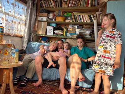 A family of four, two adults and two children, are seated on a blue sofa in a cluttered room. The room appears to be a living space with shelves full of books and various boxes stacked behind them. A window with patterned curtains is visible to the left, allowing natural light to enter the room. On a small wooden table to the left of the sofa, there are two glasses, one containing a yellow liquid, and a carton of juice. A bottle of liquid is also on the table. The people are dressed casually. The older woman on the left is wearing a pink shirt, the man next to her a black shirt, the younger woman a teal top, and the younger child a patterned dress. The younger child is holding a red lollipop. The room's decor is rustic and suggests a humble living situation. The overall impression is of a relaxed family gathering in a lived-in, possibly rural, home in Markidonovka, Russia.