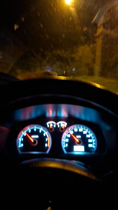 The image shows the interior of a car at night, with a focus on the illuminated dashboard. The speedometer on the right shows a speed of approximately 60 km/h, while the tachometer on the left indicates around 2,000 RPM. The windshield is visible in the background, with streetlights and foliage suggesting a location within or near a city. The overall impression is that of a car driving at night, possibly in Santan, Indonesia, given the provided context, though this specific location is not explicitly identifiable from the image itself.