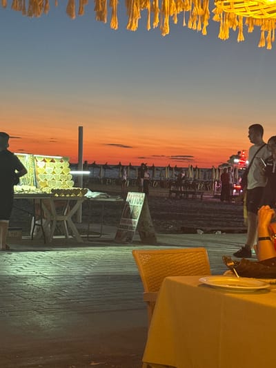 The image captures a scene at dusk on a beach in Golem, Albania. In the foreground, a table is set with a yellow tablecloth and a plate, suggesting an outdoor dining area. To the left, a vendor's stall displays numerous heart-shaped wooden items, likely souvenirs, illuminated by bright lights. A person stands behind the stall, facing away from the camera. In the background, the beach is visible with rows of closed umbrellas and lounge chairs, indicating the end of the day. Several people are scattered across the sand, some walking and others gathered near what appears to be a food stand or a kiosk. The sky displays a vibrant gradient of orange and blue, characteristic of a sunset. The overall atmosphere is relaxed and suggestive of a tourist destination.