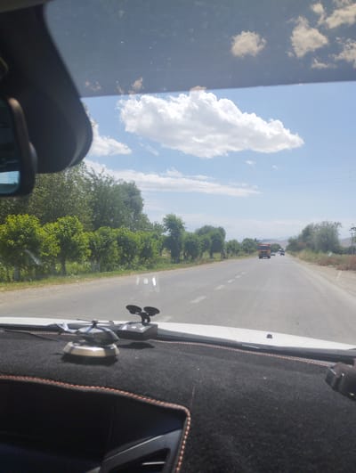 An interior view from a vehicle on a road in Egizbulak, Uzbekistan. The foreground displays a black dashboard with orange stitching, featuring a small silver helicopter figurine and a black object with two circular protrusions. Through the windshield, a two-lane asphalt road stretches into the distance under a bright blue sky with scattered white clouds. Lined with green trees and vegetation, the roadside is visible on both sides. Several vehicles, including a prominent orange truck and other cars, are traveling on the road. Electrical poles and wires are present in the background, and distant hills are discernible. The time of day appears to be daytime with clear weather conditions.