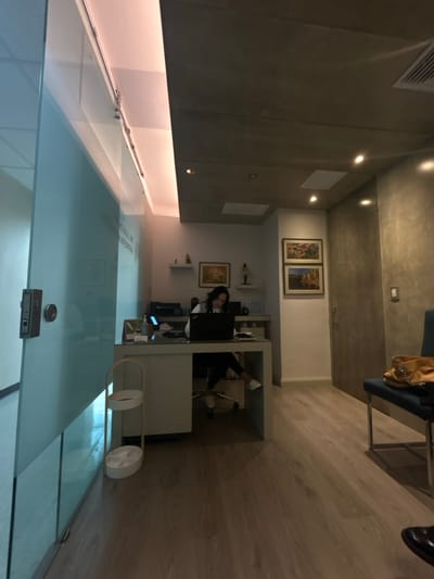 The image shows the interior of a modern office located at 9.94 latitude and -84.144 longitude in San Rafael, Costa Rica.  The scene is set in a reception area with a large glass door, a reception desk with a white countertop and a white base, and a white rolling cart. A woman is sitting in a chair behind the desk, facing a laptop, and typing. There are several framed paintings on the walls, along with a small bookshelf with objects on it. There is a second door to the right of the reception desk with a textured, gray surface. To the right of this door, a blue chair with a tan purse and other belongings is situated on the floor, with a tan handbag on the seat. The room has a light-colored wood floor and recessed lighting in the ceiling.  The overall environment is brightly lit, clean, and modern.