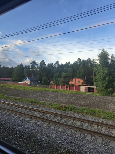 A daytime, medium shot captures a view from a train window looking out at a rural landscape near Chernaya, Russia. In the foreground, two sets of train tracks run parallel across the frame, covered in ballast and bordered by sparse green grass. Beyond the tracks, a dirt path leads to a brown wooden fence that encloses a residential area. Several houses are visible within the fenced area. One house, painted blue with a brown roof, has a distinctive blue gable with a white symbol resembling a house. Adjacent to it is a brick house with a brown roof, featuring white-framed windows and a covered garage. A dense forest of tall, dark green pine trees forms the background, with scattered deciduous trees in the foreground. The sky above is a clear blue, streaked with thin white clouds and crossed by dark power lines. The overall scene conveys a sense of peaceful, rural life on the outskirts of a Russian town.