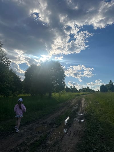 A young girl with a white cap, pink hooded sweatshirt, and striped skirt walks away from the camera on a muddy dirt road in Il'inskoe, Russia. The road is bordered by green grass and trees. The sky is partly cloudy with the sun shining through, creating lens flare. The scene appears to be set during the daytime, possibly late afternoon, given the lighting.