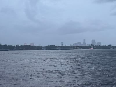 The image shows a body of water, likely a river or bay, with a cityscape in the distance. The cityscape, located in Chalmette, Louisiana, is obscured by a hazy, overcast sky, likely due to a combination of humidity and rain. The water is choppy and a dark gray color, indicating either a storm or a post-storm environment.  Several boats are visible, with some appearing to be docked.  A line of trees separates the water and the cityscape.