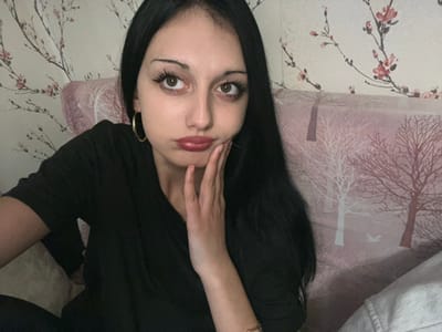 This image depicts a female individual in Lanchkhuti, Georgia, seated indoors against a patterned background. The subject has long, dark hair, brown eyes, and fair skin, with visible makeup including dark eyeliner and mascara. She wears a dark, V-neck top and a gold-colored hoop earring in her left ear, with a watch on her left wrist. Her lips are pursed, and her gaze is directed upwards and slightly to the right of the frame. Her right hand is raised, with fingers resting against the right side of her face. The background consists of two distinct patterns: to the left, a light-colored wallpaper with a cherry blossom motif, and to the right, a pink upholstered surface with a pattern of silhouetted trees and a bird.
