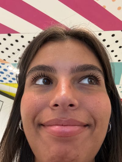 A close-up portrait features a young female subject with medium-dark skin, dark brown hair, and dark brown eyes, angled slightly upwards, occupying the majority of the frame. Her eyes are directed upwards and to the viewer's left, with eyelashes appearing enhanced by mascara. She exhibits a subtle smile with closed, glossy lips. A silver hoop earring is visible on her left ear. The background consists of brightly colored, distinct patterns, including diagonal pink and white stripes, black polka dots on white, and blue polka dots on a lighter field, consistent with an interactive art installation. This scene is located inside the Museum of Illusions at 60 N 2nd Street, Philadelphia, Pennsylvania, United States.