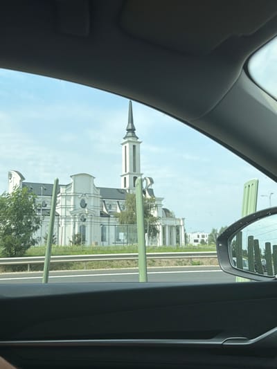 The image depicts a view from inside a vehicle looking out through the passenger side window. In the distance, a large, white, ornate building with a prominent tower topped with a spire is visible. The building appears to be a church or a similar institutional structure, characterized by its classical architectural style, symmetrical facade, arched windows, and decorative elements. The building is set against a bright blue sky with scattered clouds.
In the foreground, the interior of the car is visible, including the car's door panel and a portion of the side mirror. The side mirror reflects a row of vertical green poles and the building in the distance. The view through the window is partially obstructed by two vertical green poles that appear to be part of the road infrastructure. A highway guardrail runs horizontally across the middle ground. Trees and foliage are present to the left of the building. The overall impression is that the photo was taken while traveling on a road. The building's unique architectural style and the presence of road infrastructure suggest an urban or suburban setting. Research indicates this building is the Evangelical Lutheran Parish Church in Dybów Kolonia, Poland, also known as the "Church of the Lord's Word" or "Betlejem."