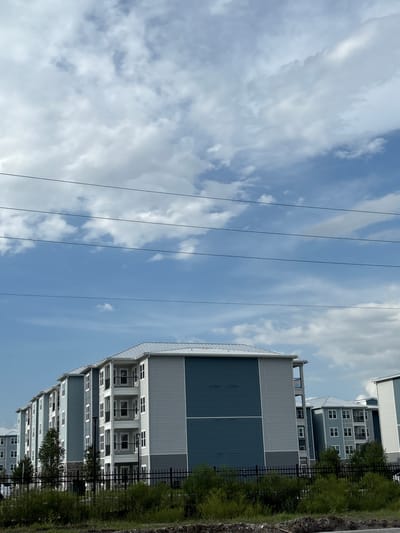 In Palmetto, Florida, United States, this image captures a view of multiple apartment buildings under a partly cloudy sky. The buildings are modern, multi-story structures with light blue and gray siding, white trim, and metal roofs. Each building features numerous balconies with black railings and white trim. In the foreground, a black metal fence runs horizontally across the frame, with green shrubbery and trees visible behind it. Overhead, several power lines are strung across the sky.
